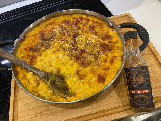 Skillet of baked macaroni and cheese with a wooden spoon and a bottle of Barbaro Mojo Best Day Ever hot sauce alongside