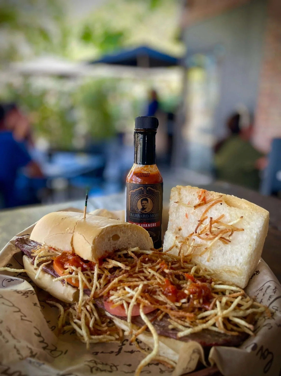 Pan con bistec on Cuban bread, layered with thin steak, tomato and a pile of shoestring potato sticks, with a bottle of Barbaro Mojo El Havanero behind it
