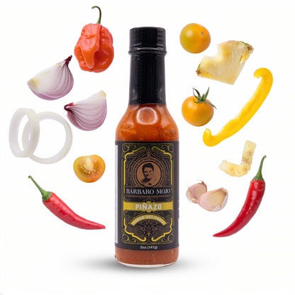Barbaro Mojo Piñazo bottle surrounded by pineapple, yellow tomatoes, habanero, Fresno peppers, yellow bell pepper, onion and garlic
